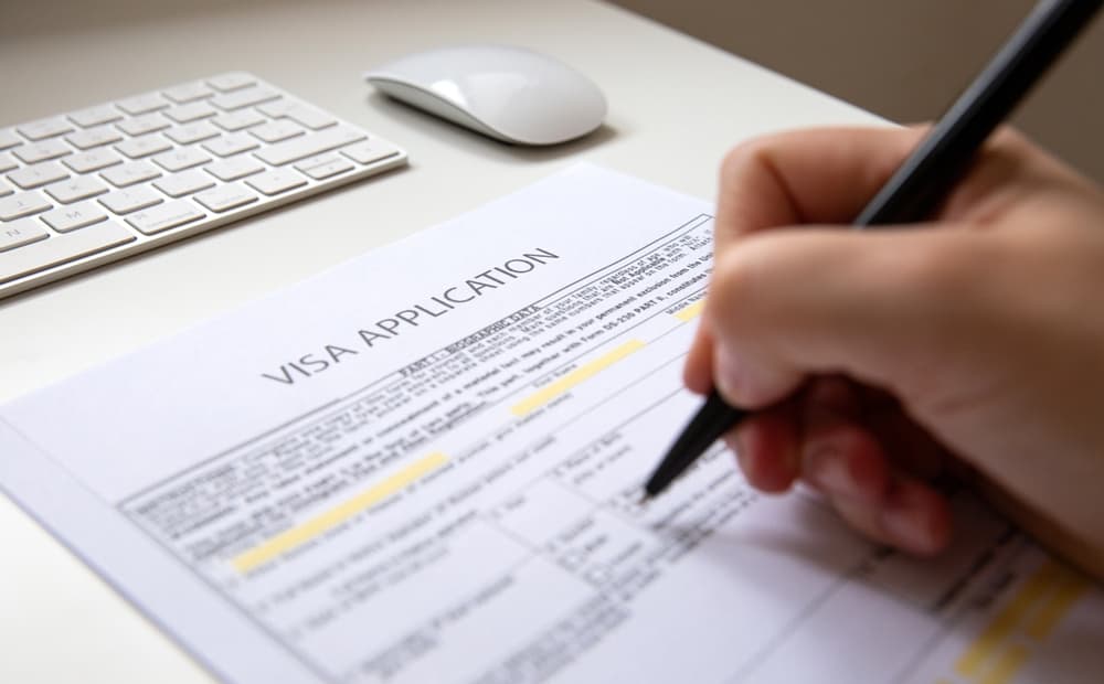 A female hand filling out a visa application document, white table with computer keyboard and a mouse in background